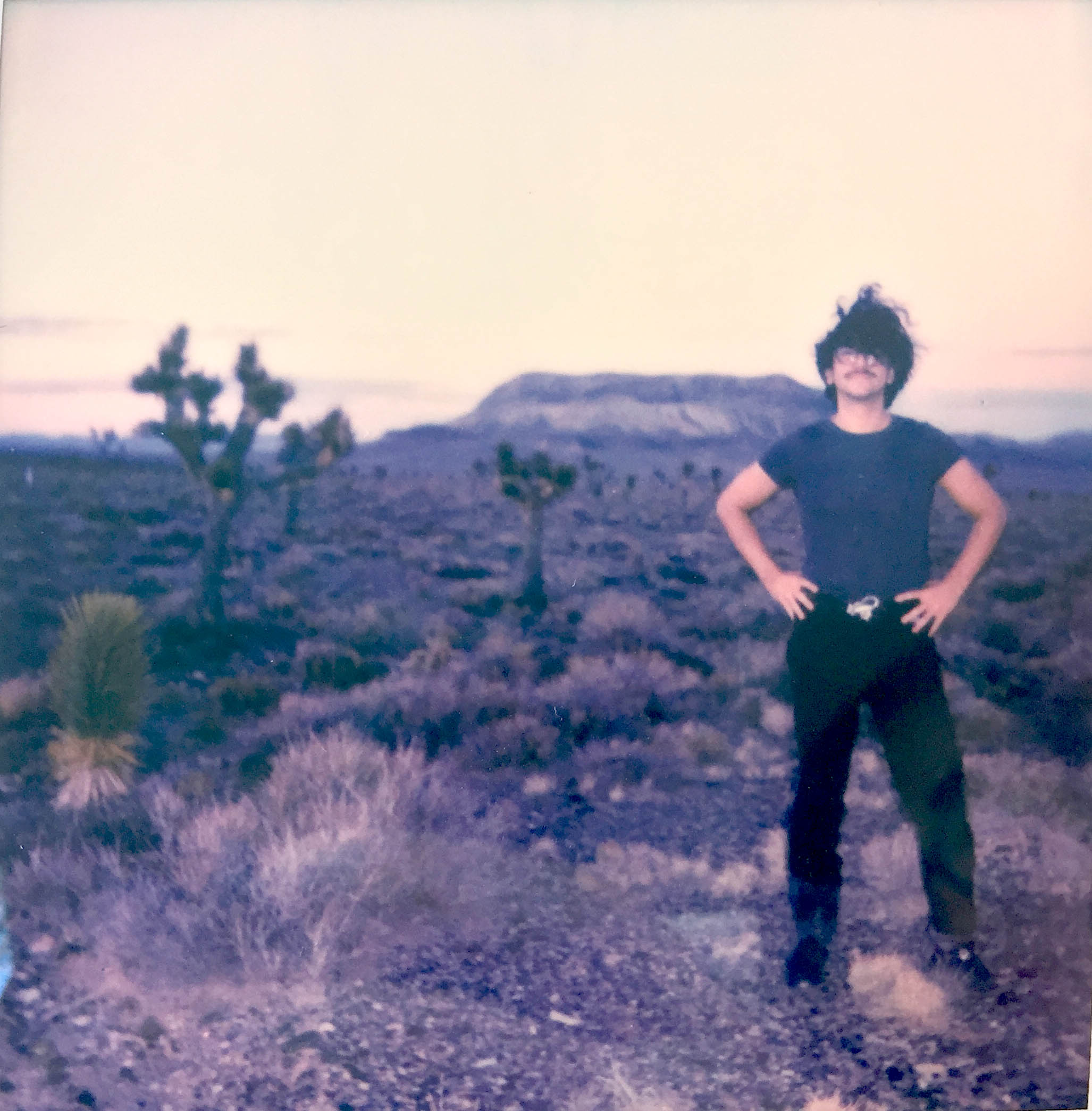 picture of an adult male in the desert. They have black hair with glasses and mustache. They are wearing a a blue tshirt, black belt/pants, and boots.